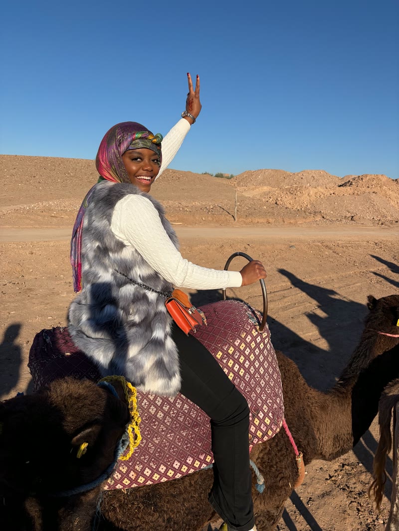Mima making a peace sign while riding a camel in the desert