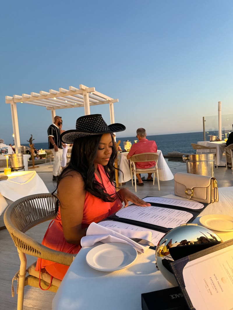 Mima in a black hat reading the menu at a seaside restaurant
